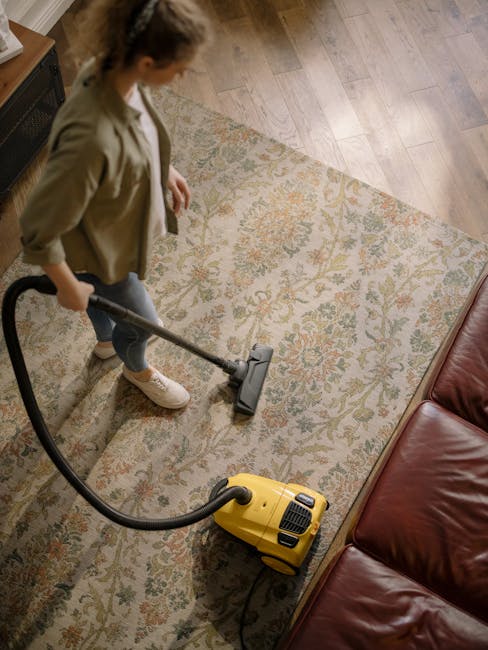 A woman using a yellow handheld vacuum cleaner to perform surface cleaning on a patterned, beige and green oriental rug in a living room. The room features a wooden floor, a red leather sofa, and a ra